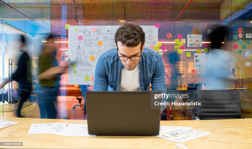 Mann, der in einem kreativen Büro mit seinem Computer arbeitet und Menschen, die sich im Hintergrund bewegen