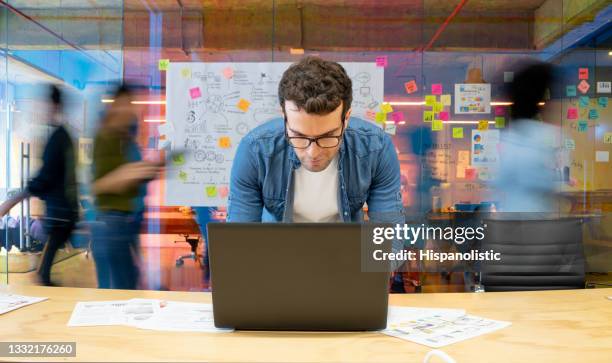 uomo che lavora in un ufficio creativo usando il suo computer e persone che si muovono in background - frenetico foto e immagini stock