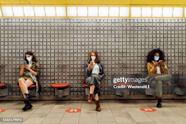 friends with protective face mask using mobile phone while sitting at social distance in subway - face mask coronavirus photos et images de collection