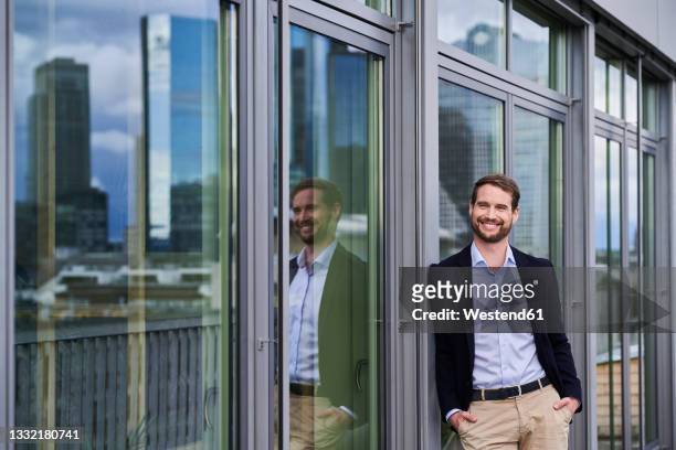 smiling male professional standing with hands in pockets leaning on glass door - office building entrance people stock pictures, royalty-free photos & images