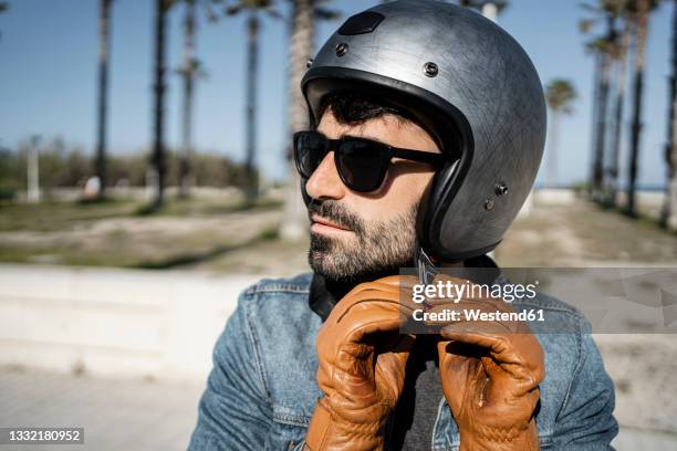 handsome man wearing glove adjusting helmet on sunny day - crash helmet stock-fotos und bilder