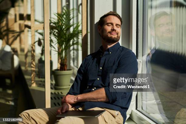 smiling male professional with eyes closed leaning on glass window at home - low key stock-fotos und bilder