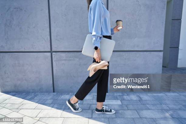 businesswoman holding laptop and high heels while walking on footpath - converse sports shoe foto e immagini stock