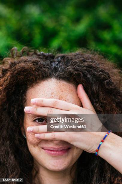 smiling curly haired woman peeking through fingers - gluren stockfoto's en -beelden