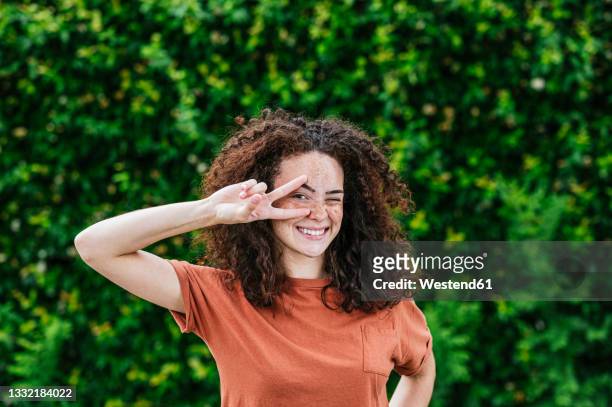 smiling young woman winking while gesturing peace sign in front of green ivy plants - oberkörper happy sommersprossen stock-fotos und bilder