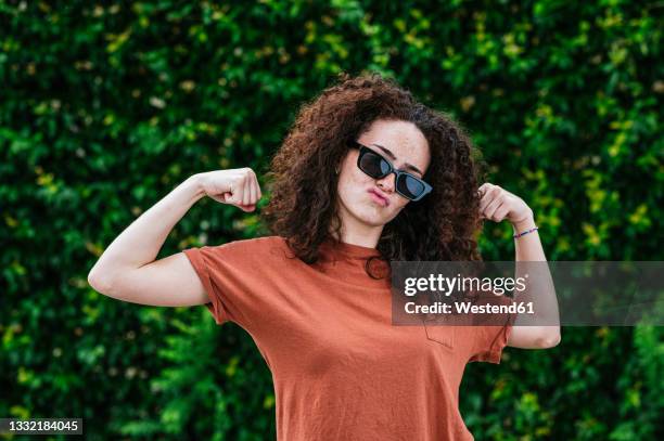 playful young woman wearing sunglasses flexing muscles in front of ivy plants - feminism stock pictures, royalty-free photos & images