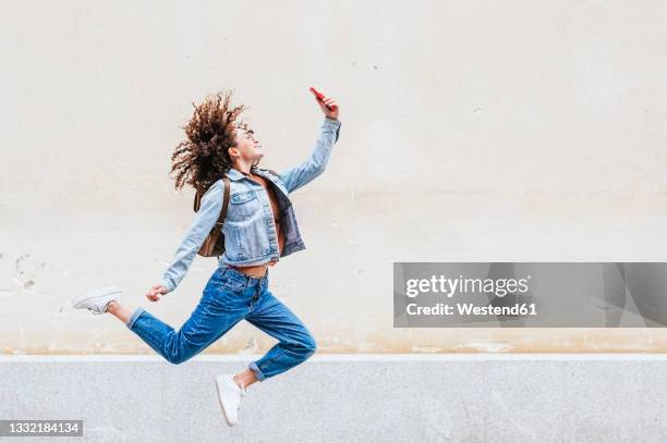excited woman taking selfie through smart phone while jumping in front of wall - happy jumping photos et images de collection