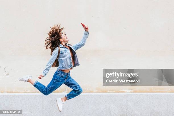 excited woman taking selfie through smart phone while jumping in front of wall - people in mid air stock pictures, royalty-free photos & images