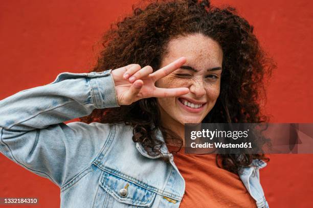 playful young woman showing peace sign while winking in front of red wall - victory sign man stock-fotos und bilder