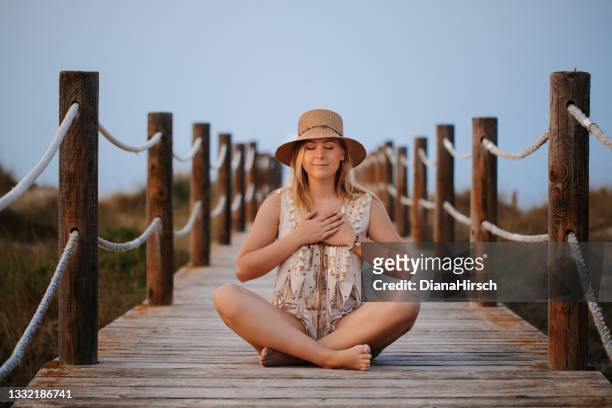 young blond spiritual woman sitting cross-legged on a wooden walkway in a mindfulness pose with her hands resting on her heart and with closed eyes with a smile - beach vibes stock pictures, royalty-free photos & images