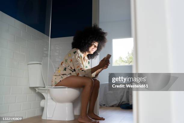 smiling woman using smart phone while sitting on toilet seat at home - woman in bathroom stockfoto's en -beelden