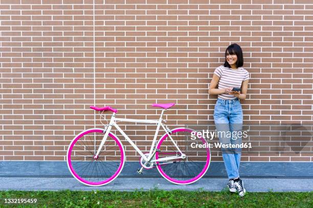 smiling young woman with smart phone leaning on wall by bicycle - phone leaning stock pictures, royalty-free photos & images