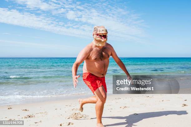 man in lizard mask running on shore at beach during sunny day - blaue shorts stock-fotos und bilder