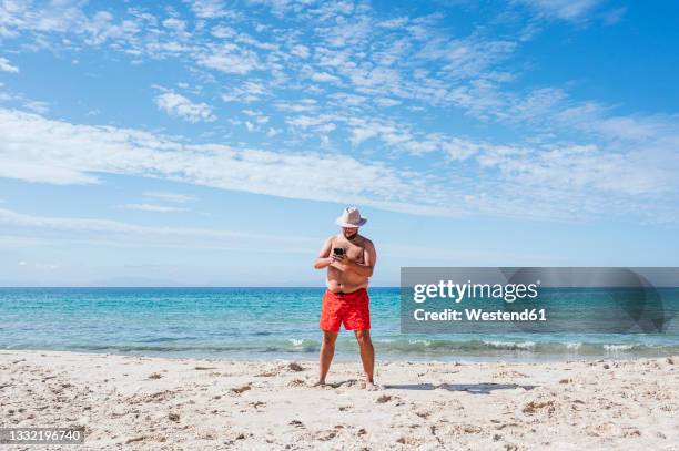 shirtless gay man using mobile phone at beach during vacation - fat guy on beach fotografías e imágenes de stock