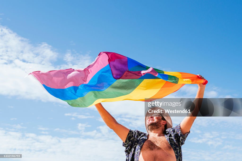 Cheerful gay man waving rainbow flag under sky on sunny day