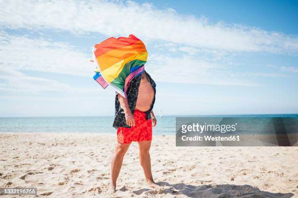 gay man with rainbow flag on face standing at beach during vacation - fat guy on beach fotografías e imágenes de stock