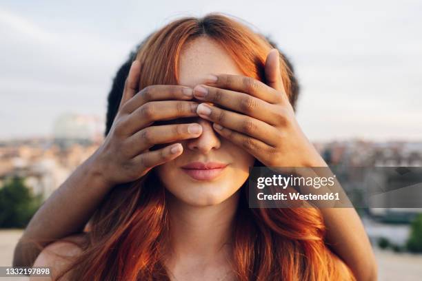 woman covering eyes of redhead girlfriend with hands at sunset - garantie stock-fotos und bilder