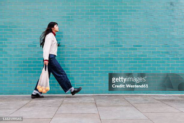 young woman with mesh bag on footpath by turquoise brick wall - gehen stock-fotos und bilder