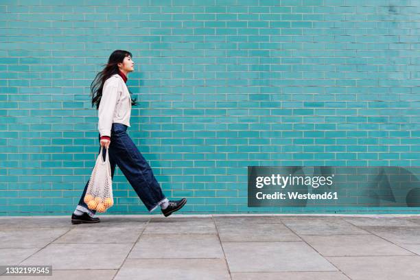 young woman with mesh bag on footpath by turquoise brick wall - marches photos et images de collection