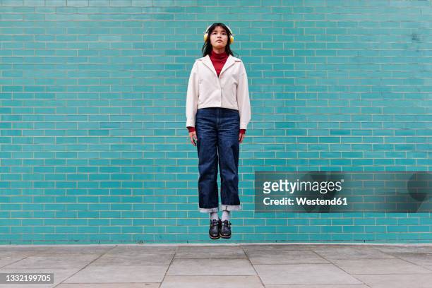 woman with headphones levitating in front of turquoise brick wall - levitación fotografías e imágenes de stock