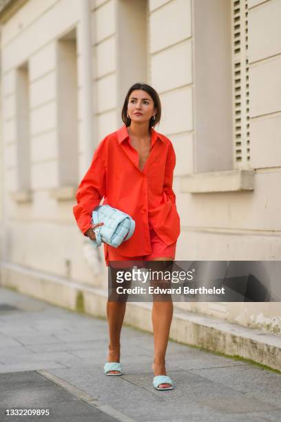 Alexandra Pereira wears earrings, a bold neon orange / red oversized shirt, matching mini shorts, a pale pastel blue woven leather bag from Bottega...