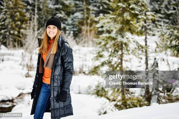 wide shot of smiling young woman standing in snow covered forest on winter afternoon - winter coat stock pictures, royalty-free photos & images