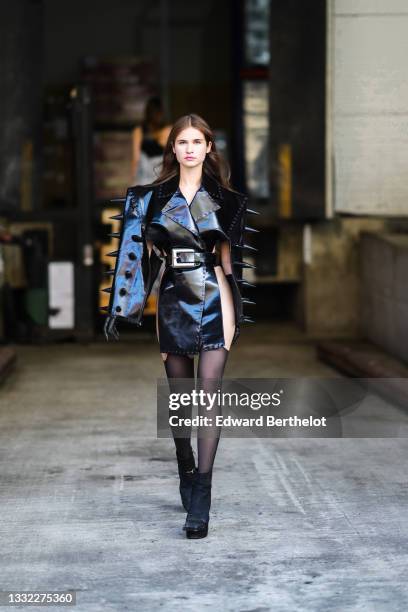 Model walks the runway and wears a black shiny leather cut-out / black large studded / epaulets / large belt dress, black tights, black suede...