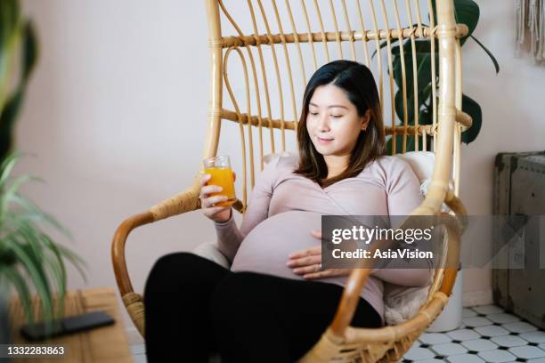 young asian pregnant woman sitting on a rattan hanging chair at home, drinking a glass of fresh orange juice while touching her belly. having a relaxing and carefree moment. wellbeing, healthy eating lifestyle during pregnancy - birthing chair stock pictures, royalty-free photos & images