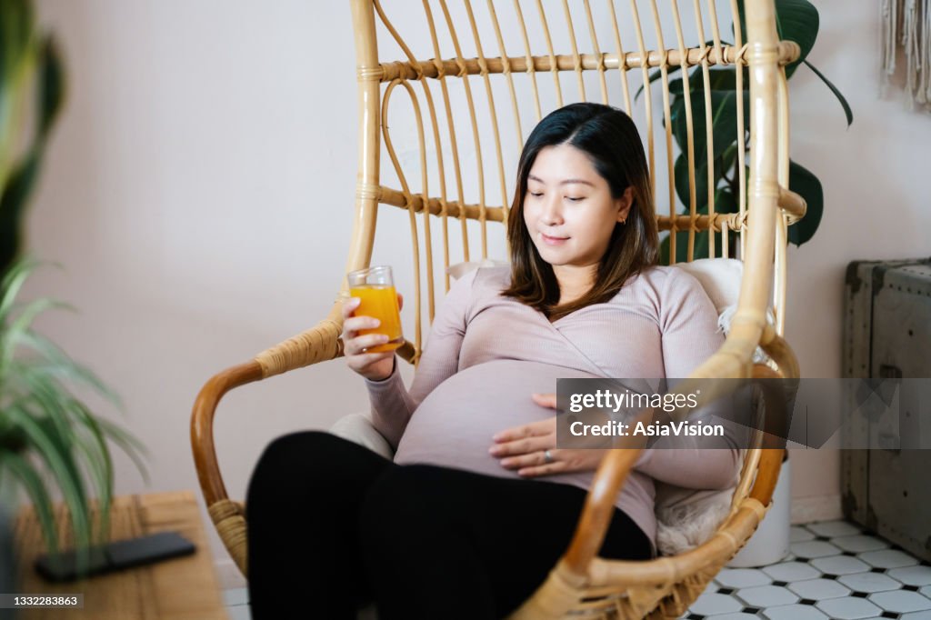 Young Asian pregnant woman sitting on a rattan hanging chair at home, drinking a glass of fresh orange juice while touching her belly. Having a relaxing and carefree moment. Wellbeing, healthy eating lifestyle during pregnancy