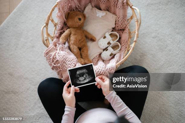 overhead view of asian pregnant woman holding an ultrasound scan photo of her baby, with a moses basket filling with baby clothing, baby shoes and soft toy teddy bear in front of her. mother-to-be. expecting a new life. preparing for the new born concept - asian woman pregnant stockfoto's en -beelden