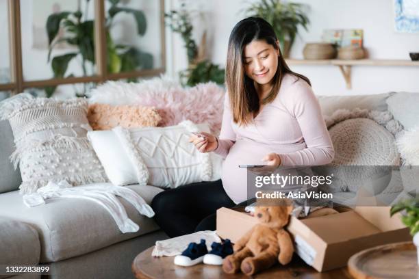 lächelnde asiatische schwangere, die online mit dem smartphone einkauft und im wohnzimmer mit kreditkarte mobil bezahlt. ein lieferpaket mit babykleidung und spielzeug auf dem couchtisch. zeit, ein paar baby-essentials für ihr ungeborenes baby zu bekomme - babybekleidung stock-fotos und bilder