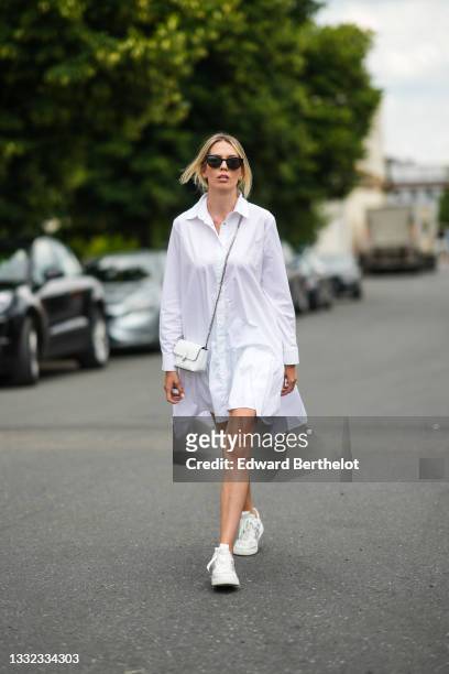 Alina Nechifor wears earrings, sunglasses, a white long ruffled shirt dress, a white shiny grained leather crossbody bag, white and gray leather...