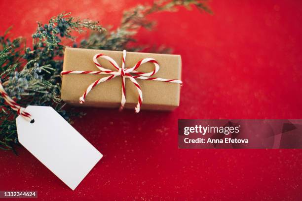 paper gift box with white tag and red-white rope bow near christmas tree branch on red table. new year coming concept. close-up. copy space for your design - gift box tag stockfoto's en -beelden