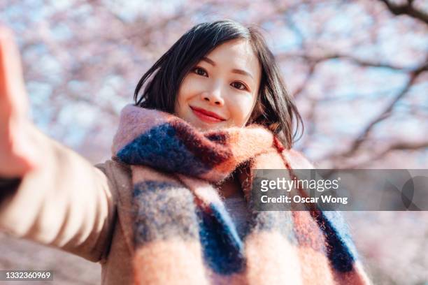 young woman taking selfie with phone under cherry blossom trees - zelfportret stockfoto's en -beelden