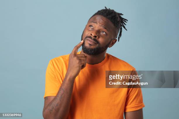 portrait of male wearing orange t-shirt looking at his chin with hand. thoughtful studio portrait against blue background - ask a question stock pictures, royalty-free photos & images