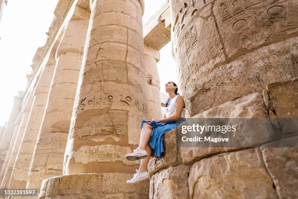 viaggiatore femminile elegante a tutta lunghezza a bassa angolazione seduto sulla costruzione in pietra e ammirando l'architettura storica nell'antico edificio storico in rovina con geroglifici su colonne intemperie - egitto foto e immagini stock