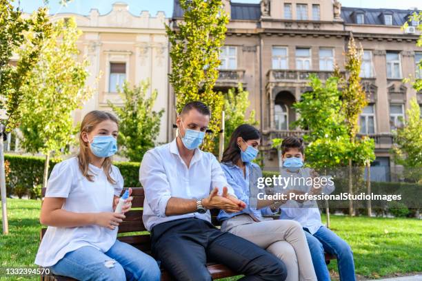 a man and woman with cute kids are sitting on the wooden bench with preventive measures due to coronavirus. - antiseptic wipe stockfoto's en -beelden