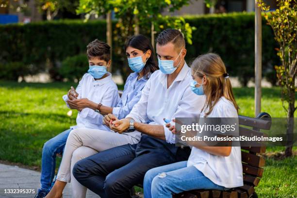 a young couple and kids with surgical medical masks is sitting on the bench and using a spray to clean his hands. - antiseptic wipe stockfoto's en -beelden