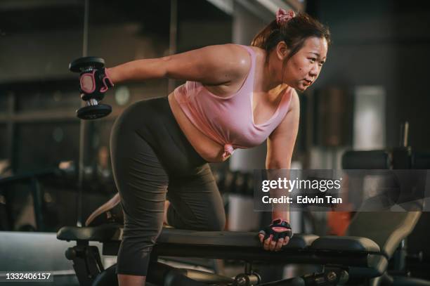 body positive asian mid adult woman exercising with dumbbells in a lunge position at gym bench at night - gewichtheffen krachttraining stockfoto's en -beelden