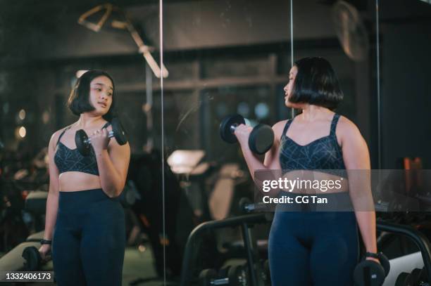 body positive asian mixed race teenage girl carrying dumbbells looking at mirror reflection in gym - body image stock pictures, royalty-free photos & images