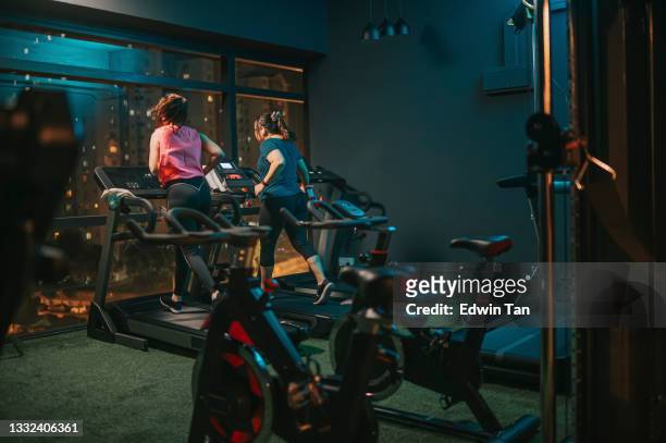 rear view body positive woman running treadmill together wiit her female friend in gym at night facing city street light - running on treadmill stock pictures, royalty-free photos & images
