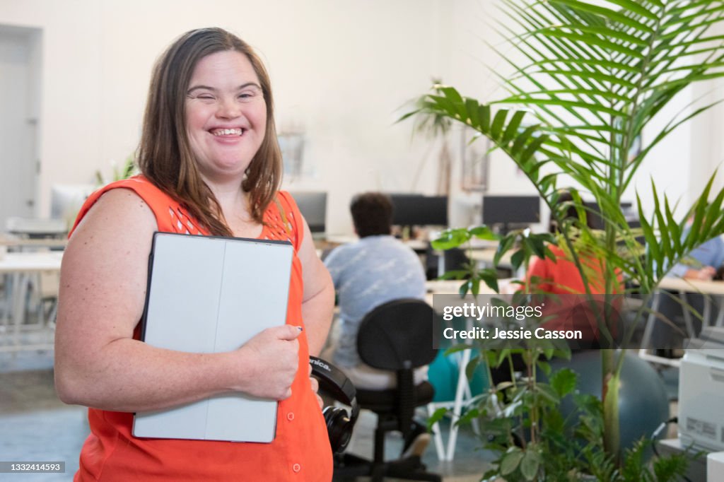 A woman with down syndrome stands in an office holding her laptop
