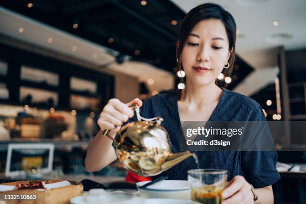 young asian woman enjoying assorted freshly served traditional dim sum in a chinese restaurant. she is pouring chinese tea in cup on the dining table. traditional chinese culture, yumcha, eating out lifestyle - asian woman drinking tea stock pictures, royalty-free photos & images