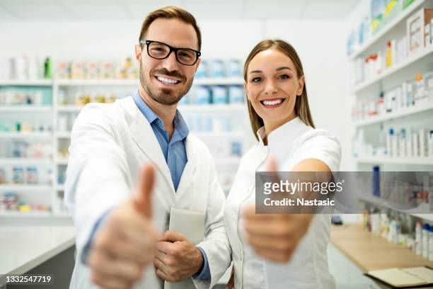 las mujeres del equipo de farmacia y el hombre en la farmacia mirando la cámara que muestra los pulgares hacia arriba - pharmacist fotografías e imágenes de stock
