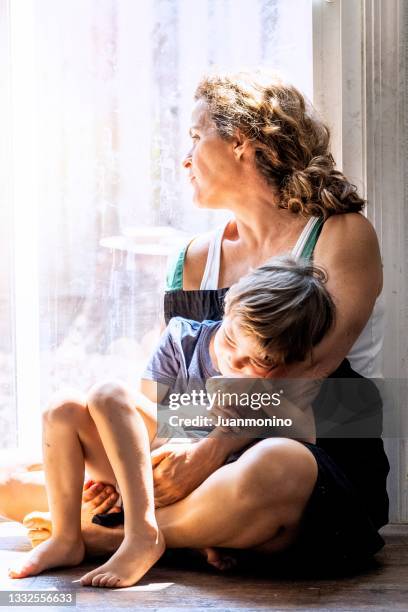 nachdenkliche reife frau, die mit ihrem sohn posiert, auf dem boden sitzt und sehr besorgt durch das fenster schaut und sich sorgen um den verlust ihres arbeitsplatzes und die räumung aufgrund der covid-19-pandemie macht - homeless child stock-fotos und bilder