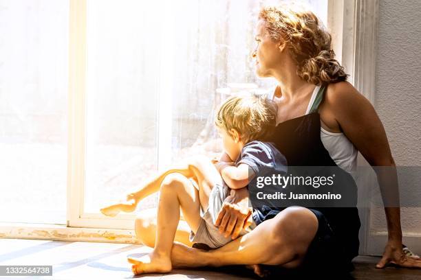 pensive mature woman posing with her son, sitting on the floor, very concerned looking through window worried about loss of her job and eviction due covid-19 pandemic - homeless woman stock pictures, royalty-free photos & images
