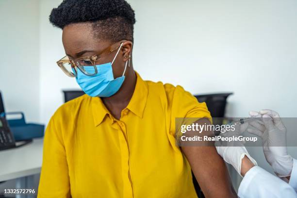 young african american woman getting flu shot during seasonal vaccination campaign. doctor or nurse in medical face mask cleans skin on patient's arm before injecting modern covid 19 antiviral vaccine - immunology stock pictures, royalty-free photos & images