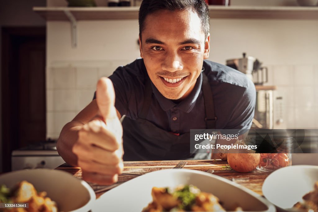 Porträt eines jungen Mannes, der Daumen hoch zeigt, während er zu Hause eine köstliche Mahlzeit zubereitet