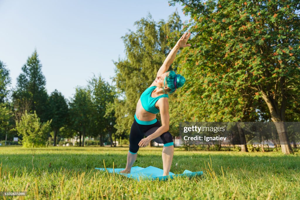 Attractive beautiful young woman with a turquoise hair in a turquoise yoga outfit doing yoga on a grass in a park, during sunrise or sunset, stretching to the side to the bent leg on a yoga mat, enjoying her practice