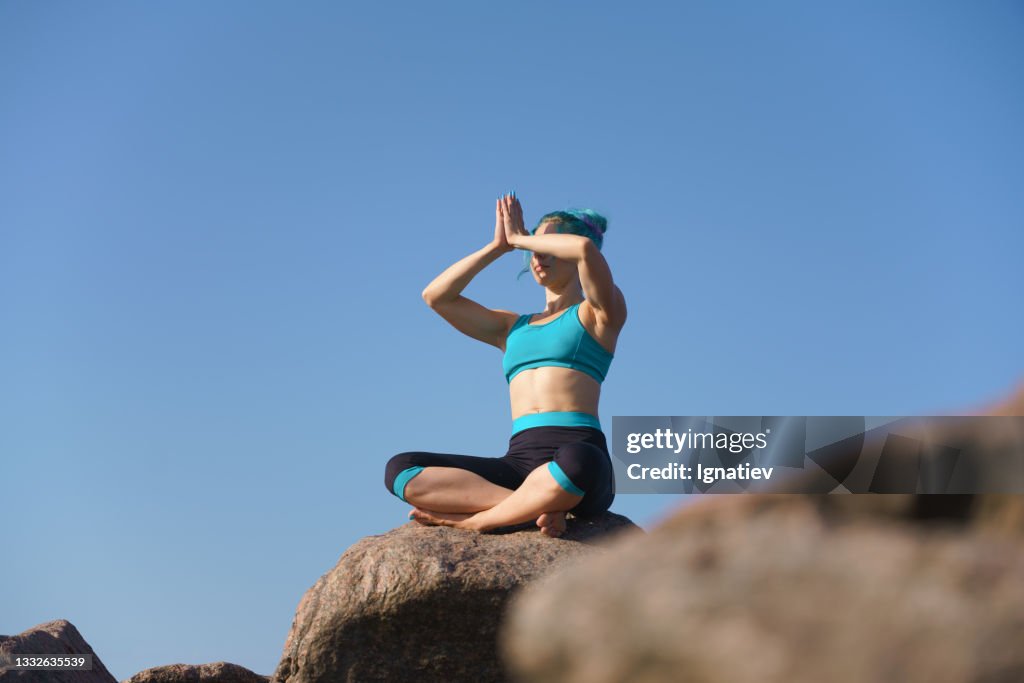 Attractive Beautiful Fit Young Woman With A Turquoise Hair In A  Turquoiseblack Yoga Outfit Sits On A Stone Against The Sky With Her Hands  In Front Of Her Face Palms Together High-Res